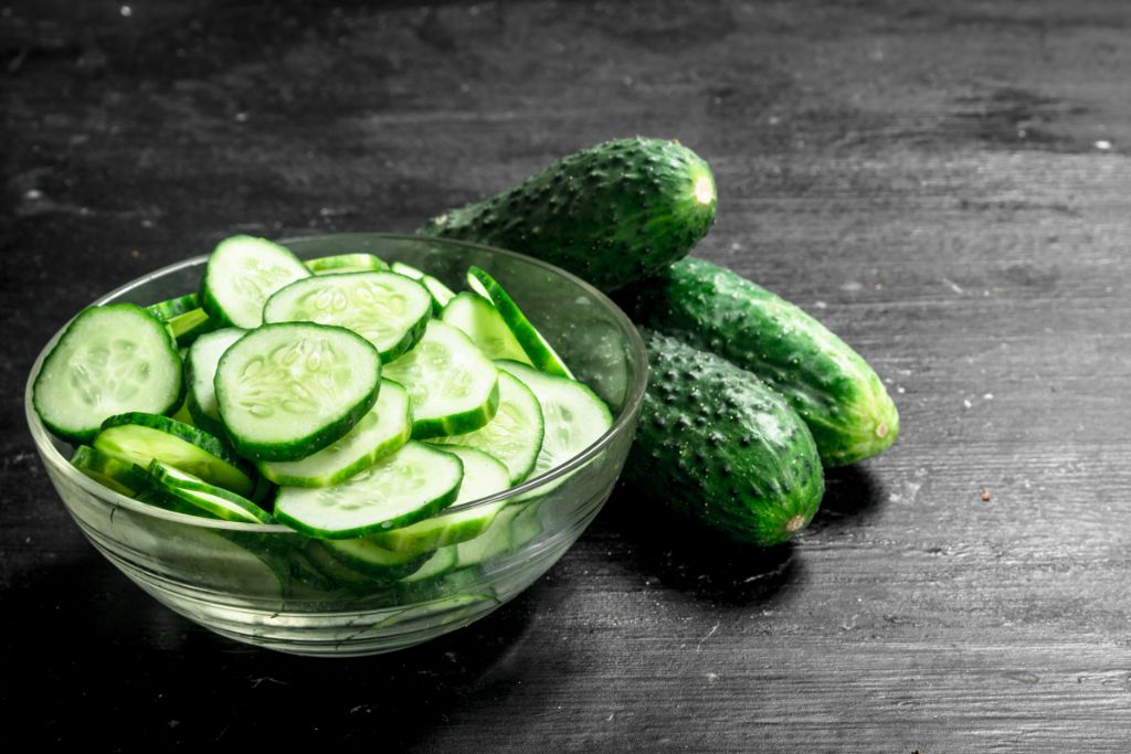 sliced cucumber in a glass bowl beside three whole cucumber placed on a dark table
